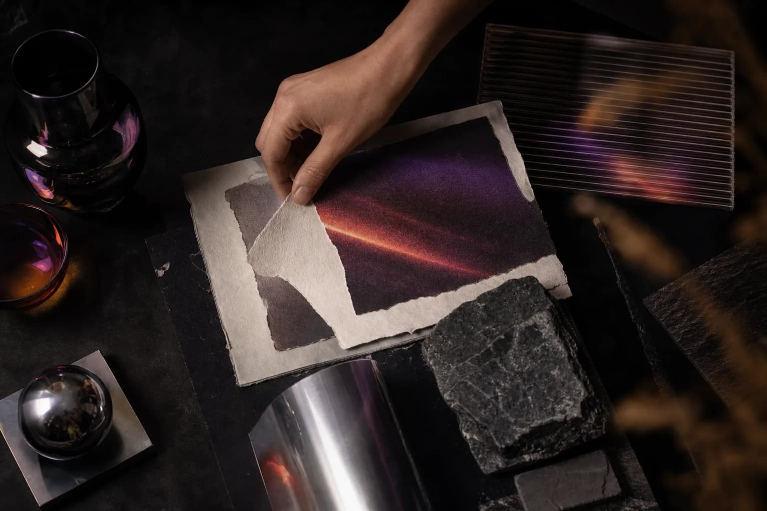 A hand arranging torn art prints among glass, metal, and raw stone
